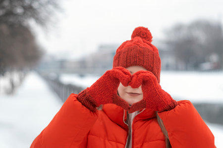 Portrait of a beautiful girl in a red jacket and hat shows heart sign with her palms on a background of a winter park.の写真素材