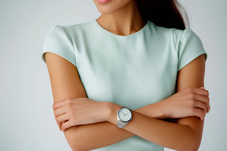 Businesswoman in a light green dress confidently displaying her wristwatch while posing with folded arms against a gray backgroundの素材