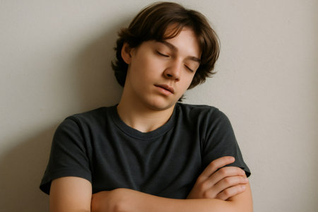 Sad young man with closed eyes and crossed arms is leaning against white wall, suffering from loneliness and depressionの素材