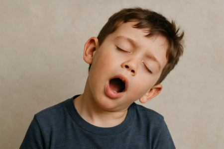 Young boy yawning with his eyes closed, expressing tiredness or boredom against a neutral backgroundの素材