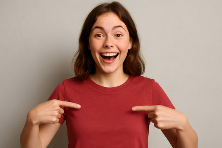 Young woman pointing at herself with an amazed and joyful expression, radiating happiness against a soft gray backgroundの素材
