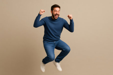 Young man with beard and casual clothes jumping with raised fists, expressing joy and excitement on a neutral backgroundの素材