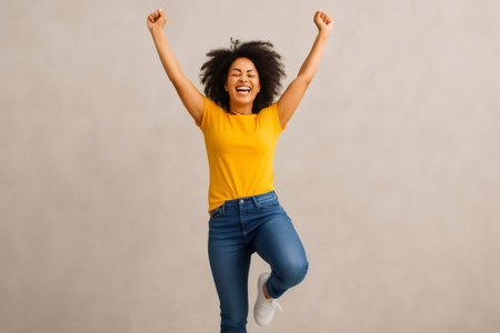 Young woman with curly hair and yellow t shirt joyfully leaping and celebrating success with arms raisedの素材