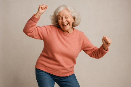 Elderly woman with gray hair and pink sweater dancing and smiling, celebrating victory or achievement on neutral backgroundの素材