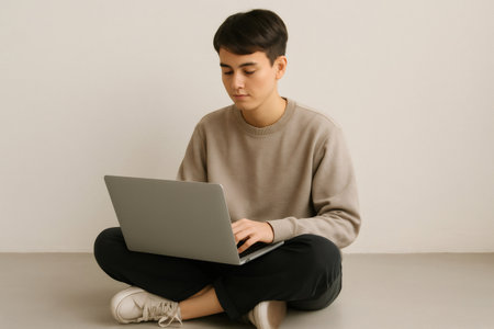 Focused young man sitting cross legged on the floor, working intently on his laptop while immersed in the digital world of online tasksの素材