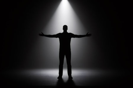 Silhouette of confident man standing with open arms under spotlight in dark room, embracing success and new opportunitiesの素材