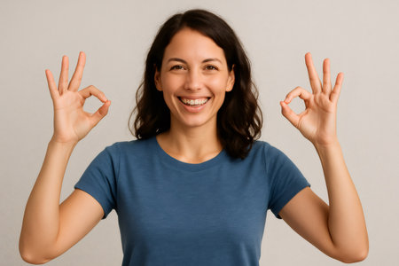 Portrait of a young woman smiling brightly while making an ok gesture with both hands, radiating positivity against a light backgroundの素材