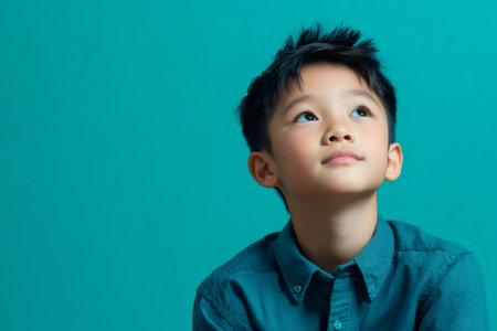 Portrait of a pensive young boy looking up while wearing a blue shirt, isolated on a turquoise backgroundの素材