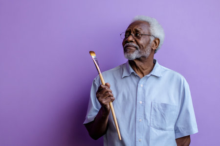 Pensive senior african american artist holding paintbrush and looking up against vibrant purple backdropの素材