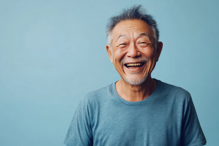 Happy senior Asian man with gray hair and a goatee laughing joyfully against a vibrant blue background, radiating positivity and cheerの素材