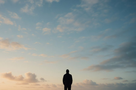 Silhouette of a man standing under a vast sky with scattered clouds during a breathtaking sunset, conveying a sense of peace and tranquilityの素材