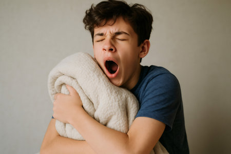 Young man waking up and yawning while wrapped in a cozy blanket, feeling the weight of sleepiness and tiredness in the morning lightの素材