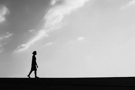 Black and white image highlighting the minimalist silhouette of a person walking against a clear skyの素材