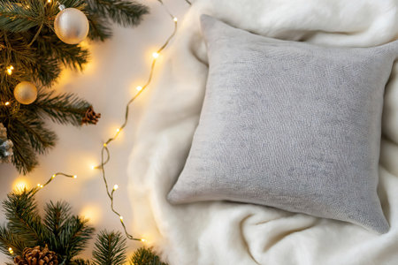 Gray blank pillow mockup is lying on a white blanket near a Christmas tree decorated with silver baubles and a garlandの素材