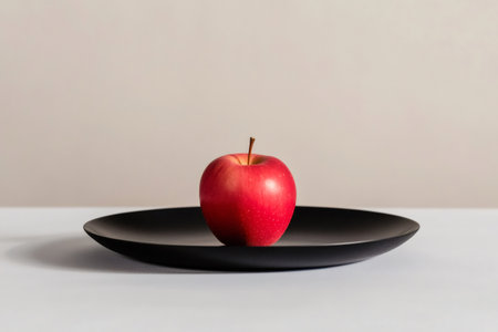 Vibrant red apple sits on a sleek, black plate, creating a striking contrast against a muted background in a minimalist still life compositionの素材