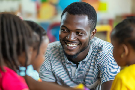 Happy male teacher interacting with two young girls in a colorful preschool classroom, promoting early childhood education and developmentの素材