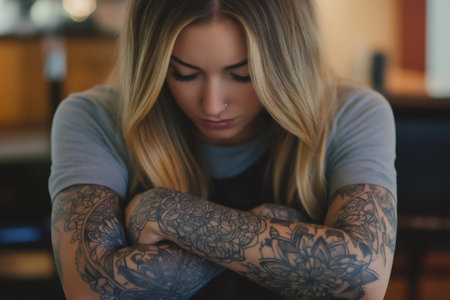 Tattoo artist with crossed arms and floral tattoos looking down, showing a serious and focused expressionの素材