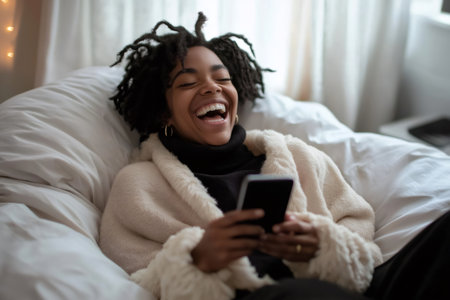 Cheerful young woman is using her smartphone while relaxing on a comfortable beanbag chair in her cozy bedroomの素材