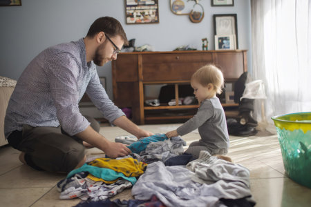 Stay at home dad folding laundry while entertaining in playful moments with his toddler son in a cozy bedroom settingの素材