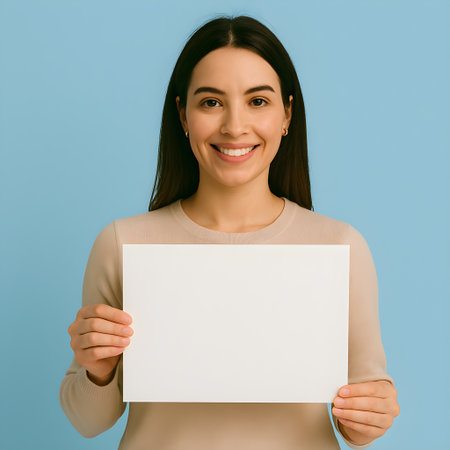 Smiling woman showing blank white paper sheet with copy space for your text or image, isolated over blue backgroundの素材