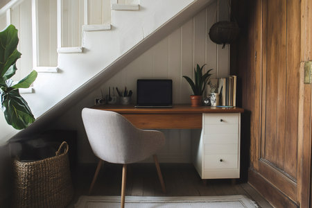 Cozy and functional workspace located under a staircase, featuring a laptop, plants, and organizational elements, creating an inspiring and productive environmentの素材