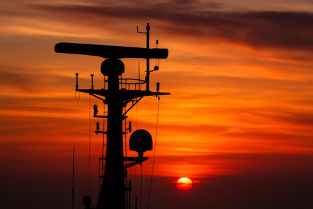 Ship radar mast silhouetted against a vibrant orange sunset, creating a dramatic maritime sceneの素材