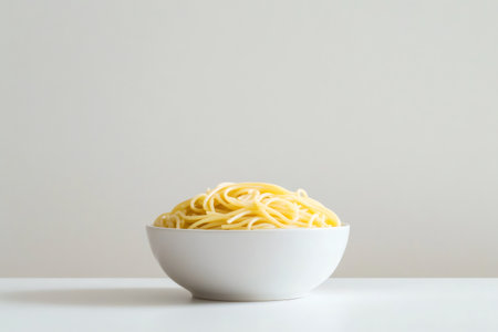 Simple white bowl filled with a mound of cooked spaghetti sits on a white table against a neutral background, creating a minimalist food imageの素材