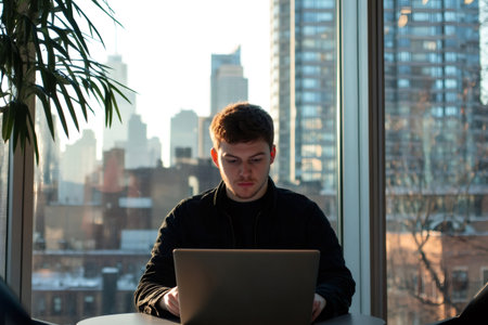 Young man concentrating on his computer, working remotely or studying with a modern city skyline and bright window in the backgroundの素材
