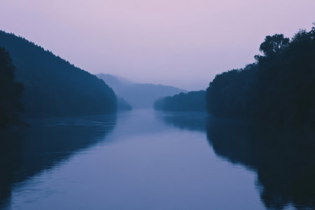 Calm river flowing between dark forest hills under a soft purple sky, creating a tranquil and atmospheric landscapeの素材