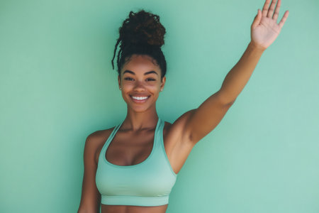 Young African American woman in athletic wear greeting, smiling and waving hand, expressing positivity and warmthの素材