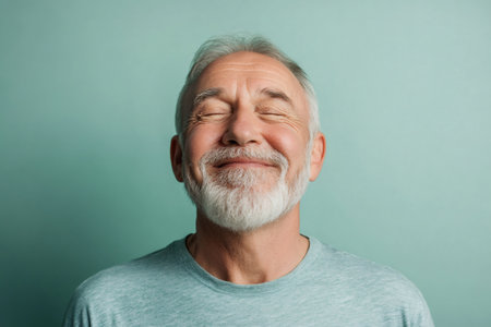 Older man with a white beard and gray hair smiling peacefully with closed eyes against a solid teal backgroundの素材
