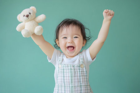 Cheerful infant baby expressing joy and excitement, raising hands with a toy bear and clenched fist against a plain backgroundの素材