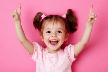 Happy child with pigtails pointing fingers up, expressing joy and excitement against a pink backgroundの素材