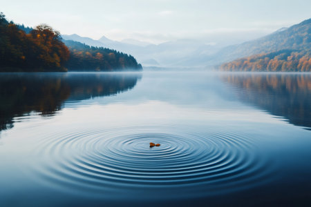 Autumn leaf dropping into serene lake water, sending concentric ripples across the surface with misty mountains and fall treesの素材