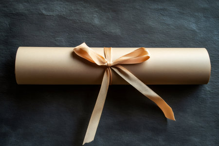 Rolled brown paper scroll, secured with a golden ribbon, lying on a dark textured surface. Representing diploma, invitation, or awardの素材