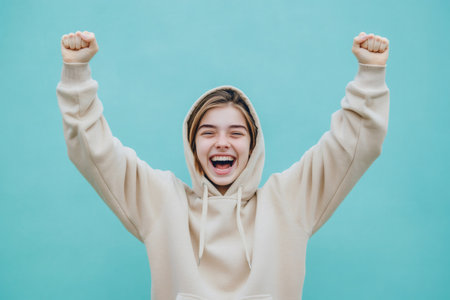Teenager wearing a hoodie, raising both arms with clenched fists, feeling joyful, celebrating triumph and victoryの素材