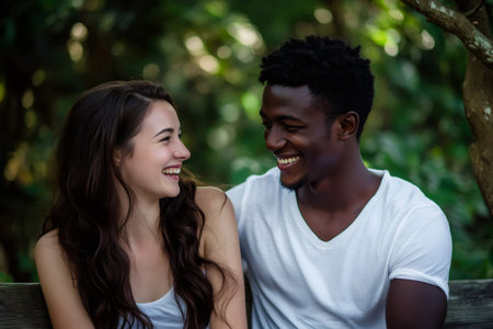 Young man and woman with different skin tones smiling and laughing together, sitting on a bench in a natural settingの素材