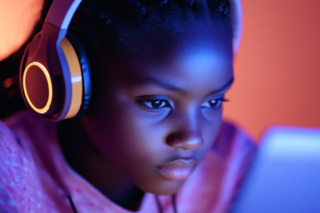 Young person wearing headphones with illuminated earcup, intensely focusing on a screen with blue light reflecting on their faceの素材