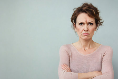 Woman showing a serious, displeased expression with furrowed brows, pursed lips, and crossed arms on a simple backgroundの素材