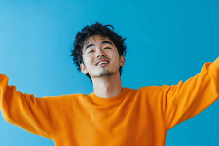 Young Asian man feeling happy and healthy, wearing a casual orange sweater with arms spread wide on a blue backgroundの素材