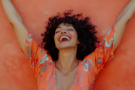 Young woman expressing pure joy and freedom, laughing with eyes closed while raising her arms against an orange backgroundの素材