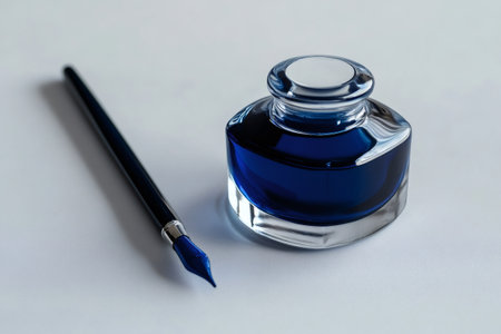 Glass inkwell filled with vibrant blue ink, sitting beside a traditional dip pen on a clean white surfaceの素材