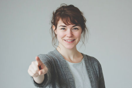 Young woman looking at viewer, smiling and pointing an index finger forward. Engaging person creating a connectionの素材