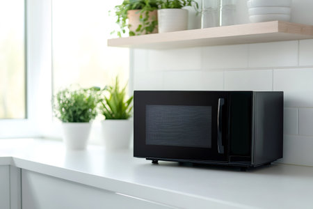 Sleek black microwave appliance sitting on a white kitchen countertop, with green plants and a wooden shelf in a bright backgroundの素材