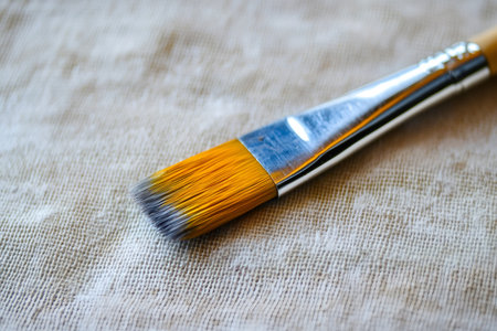 Paintbrush showing yellow bristles with dried gray paint, lying on a textured light beige canvas surfaceの素材