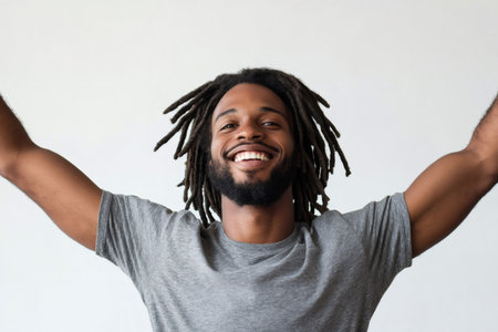 Young Black man with dreadlocks smiling brightly, raising arms in success and celebration. Expressing happiness, freedom, and positive emotions on a plain backgroundの素材