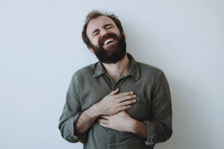 Bearded man bending over and holding chest laughing out loud against a white background, representing humor and good moodの素材