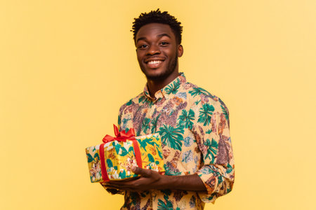 Young African American man holding a colorful wrapped gift box with a red ribbon, happily anticipating a celebration or surpriseの素材