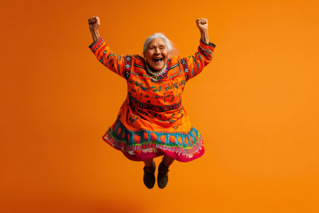 Lively Indigenous elder woman jumping high with arms raised, expressing joy and freedom on an orange backgroundの素材