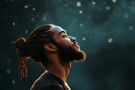African American man looking up with closed eyes, feeling peace and hope, illuminated by golden light against bokeh backgroundの素材